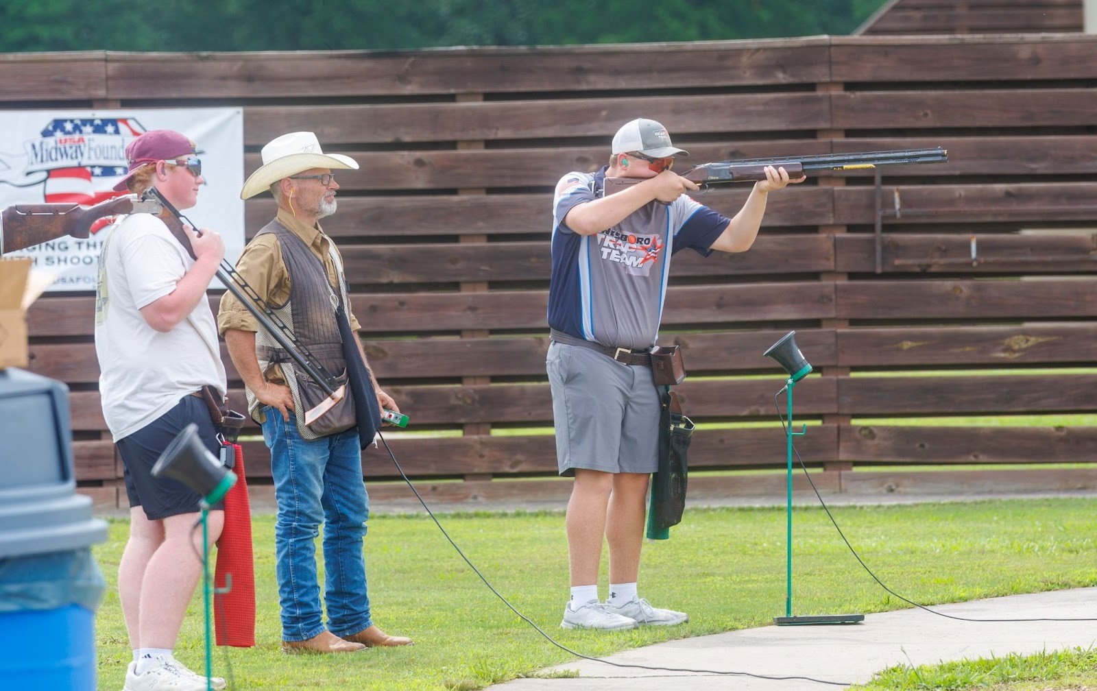 Shooter aims a shotgun at an outdoor range while two men in hats watch nearby. A wooden fence is in the background.