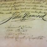 Close-up of an old parchment with flowing cursive signatures, prominently featuring the signature 'John Hancock'.