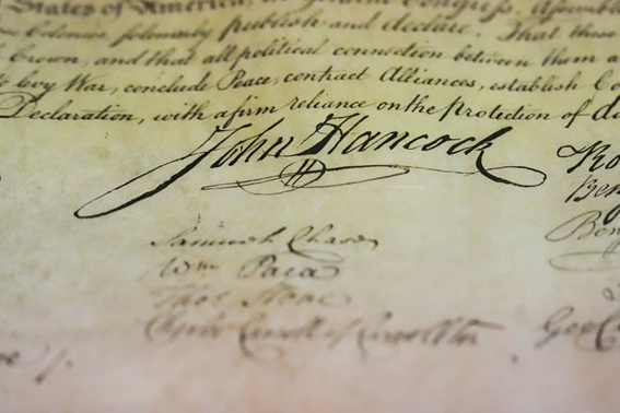 Close-up of an old parchment with flowing cursive signatures, prominently featuring the signature 'John Hancock'.