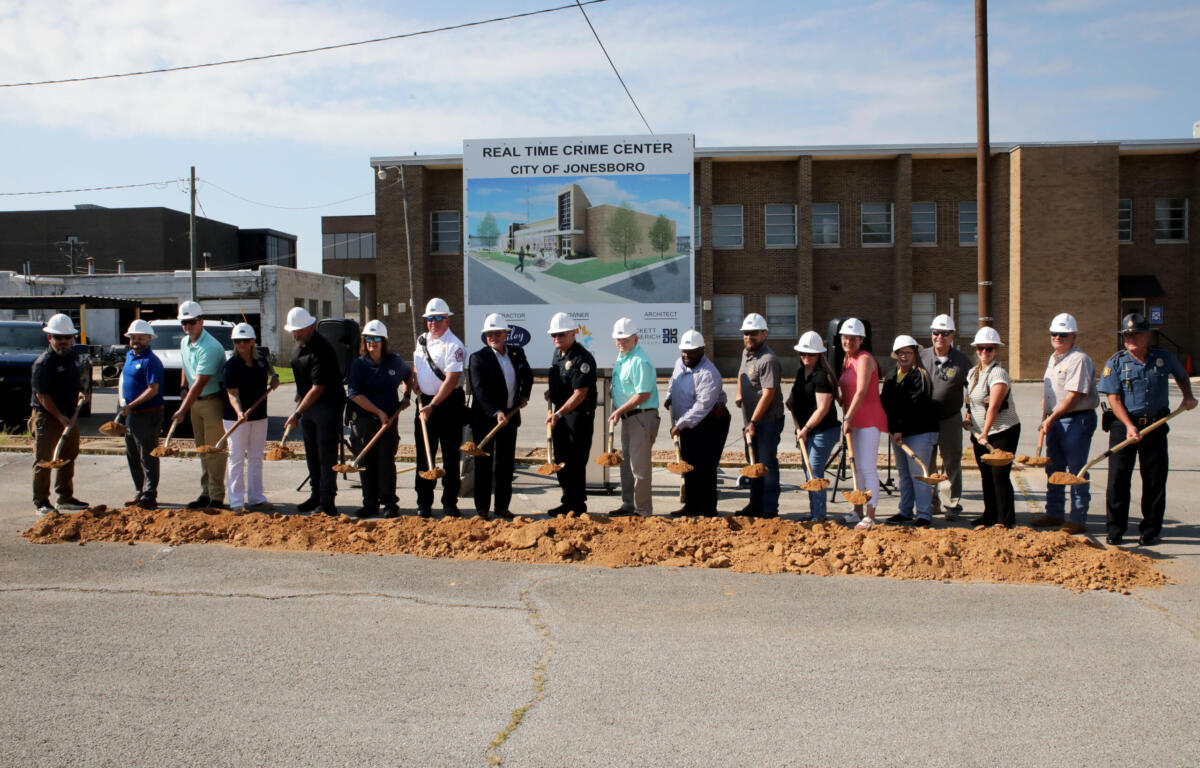 People in hard hats stand in a line with shovels at a groundbreaking ceremony in front of a sign for the Real Time Crime Center, City of Jonesboro, and a brick building behind them.