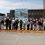 People in hard hats stand in a line with shovels at a groundbreaking ceremony in front of a sign for the Real Time Crime Center, City of Jonesboro, and a brick building behind them.