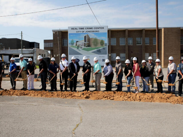 People in hard hats stand in a line with shovels at a groundbreaking ceremony in front of a sign for the Real Time Crime Center, City of Jonesboro, and a brick building behind them.