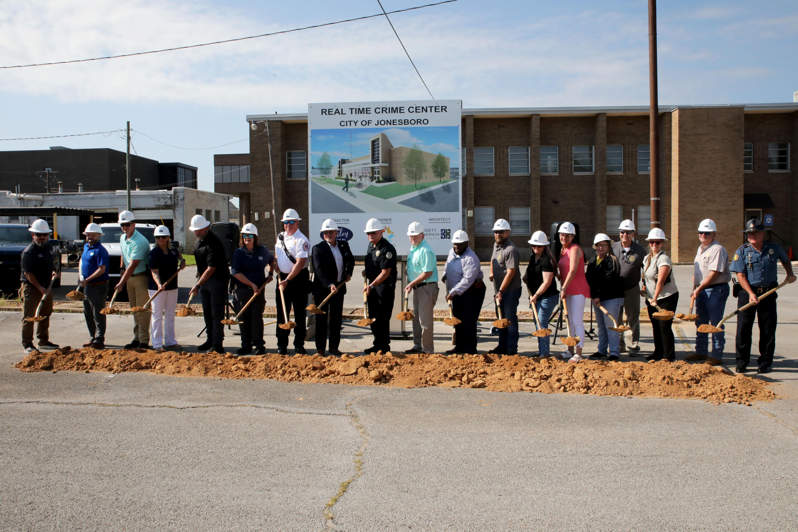 People in hard hats stand in a line with shovels at a groundbreaking ceremony in front of a sign for the Real Time Crime Center, City of Jonesboro, and a brick building behind them.