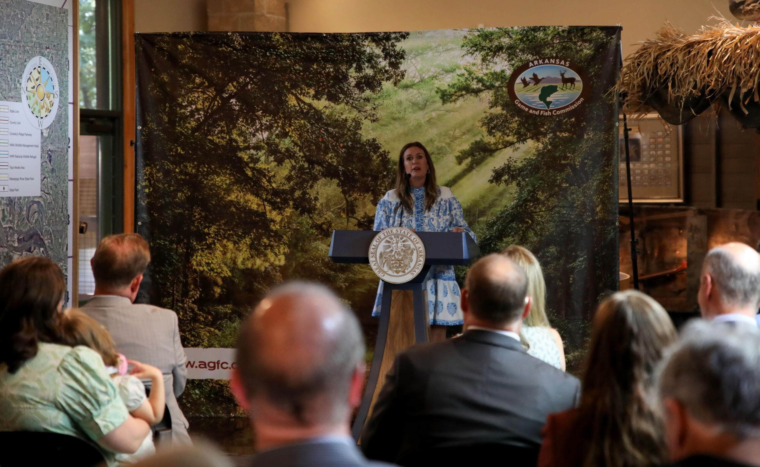 Woman in a blue patterned dress speaks at a podium, with a woodland backdrop and an audience seated in front of her, Arkansas seal visible on the podium emblem.