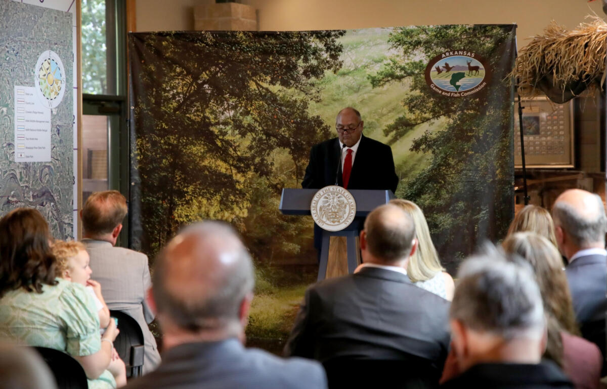 Man in suit and red tie speaks at a lectern with state seal, audience listening, backdrop shows trees and Arkansas Game and Fish Commission logo.