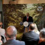 Man in suit and red tie speaks at a lectern with state seal, audience listening, backdrop shows trees and Arkansas Game and Fish Commission logo.