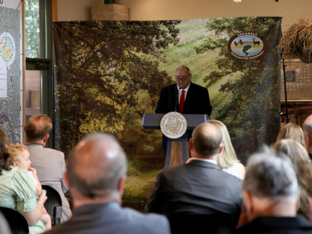 Man in suit and red tie speaks at a lectern with state seal, audience listening, backdrop shows trees and Arkansas Game and Fish Commission logo.
