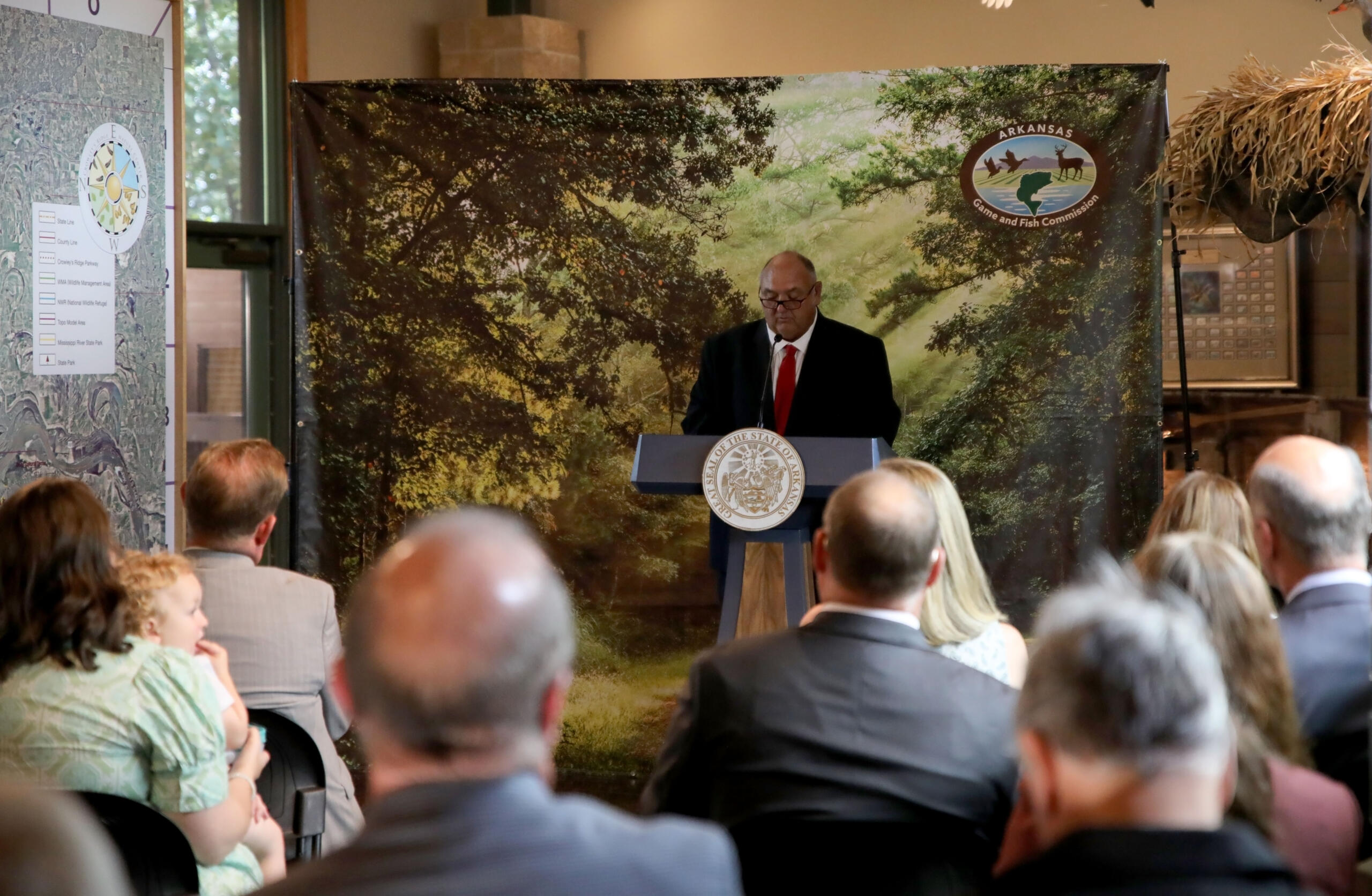 Man in suit and red tie speaks at a lectern with state seal, audience listening, backdrop shows trees and Arkansas Game and Fish Commission logo.