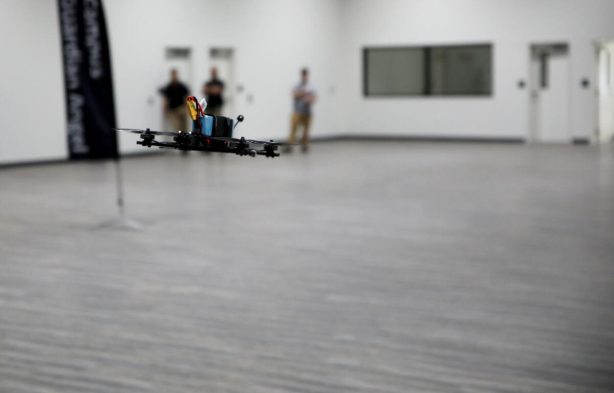 Quadcopter drone hovers low over a gray gym floor indoors, with three blurred onlookers in the background.