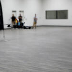 Quadcopter drone hovers low over a gray gym floor indoors, with three blurred onlookers in the background.
