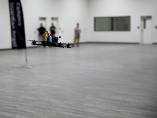 Quadcopter drone hovers low over a gray gym floor indoors, with three blurred onlookers in the background.