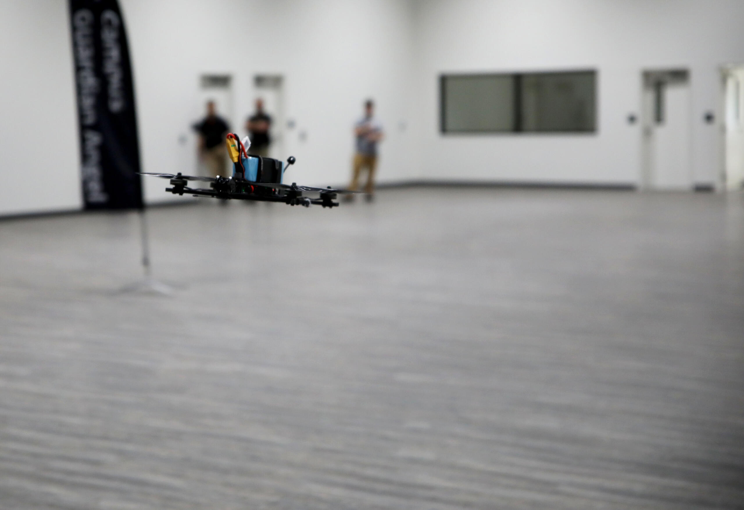 Quadcopter drone hovers low over a gray gym floor indoors, with three blurred onlookers in the background.