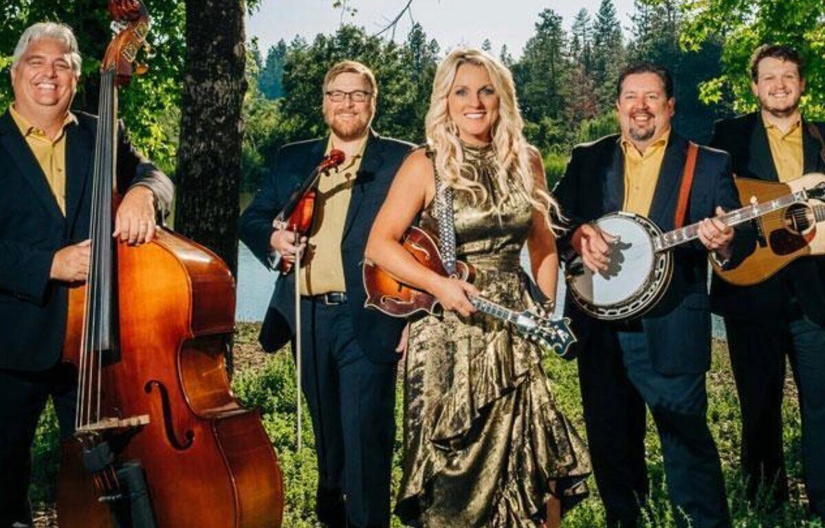 Five-member band outdoors: a woman in a metallic gold dress holding a violin, flanked by four men with a upright bass, violin, banjo, and guitar.