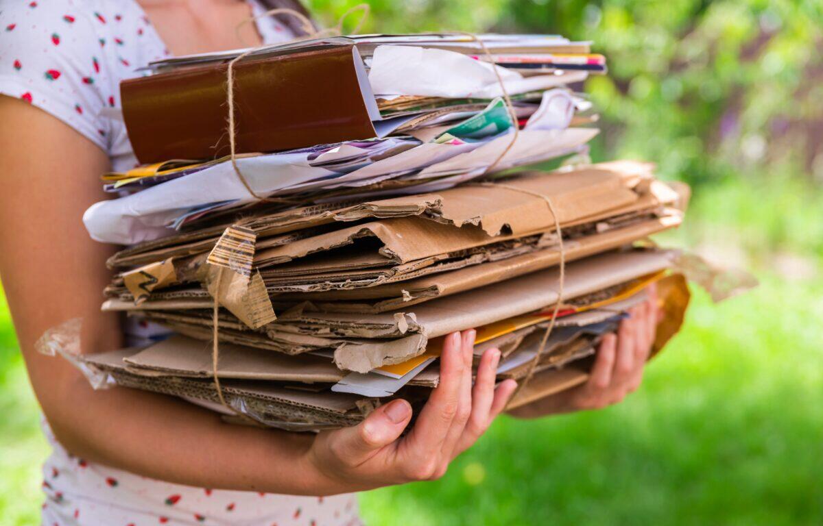 Person wearing a white dotted dress holds a large bundled stack of cardboard, papers, and envelopes tied with string outside.