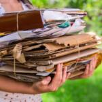 Person wearing a white dotted dress holds a large bundled stack of cardboard, papers, and envelopes tied with string outside.