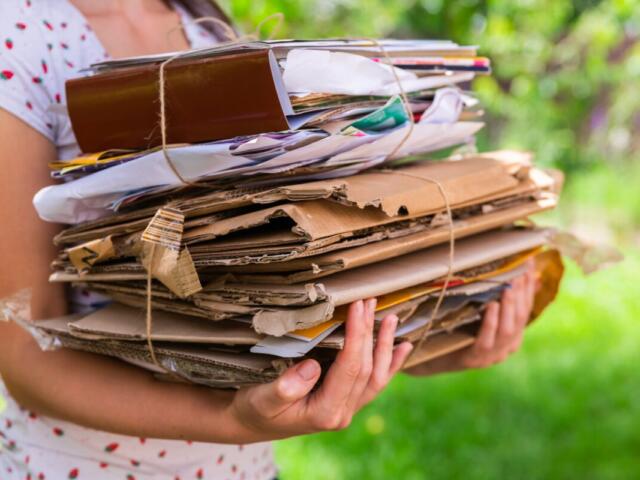 Person wearing a white dotted dress holds a large bundled stack of cardboard, papers, and envelopes tied with string outside.
