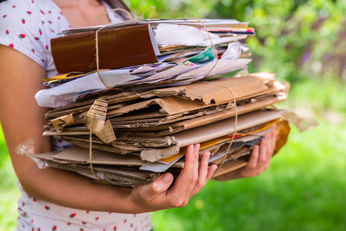Person wearing a white dotted dress holds a large bundled stack of cardboard, papers, and envelopes tied with string outside.