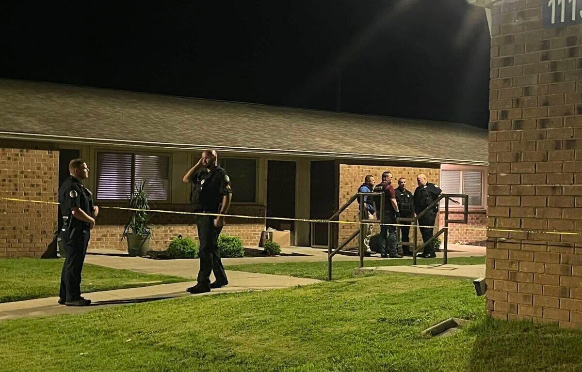 Police officers stand behind yellow caution tape outside a brick apartment building at night, investigating.