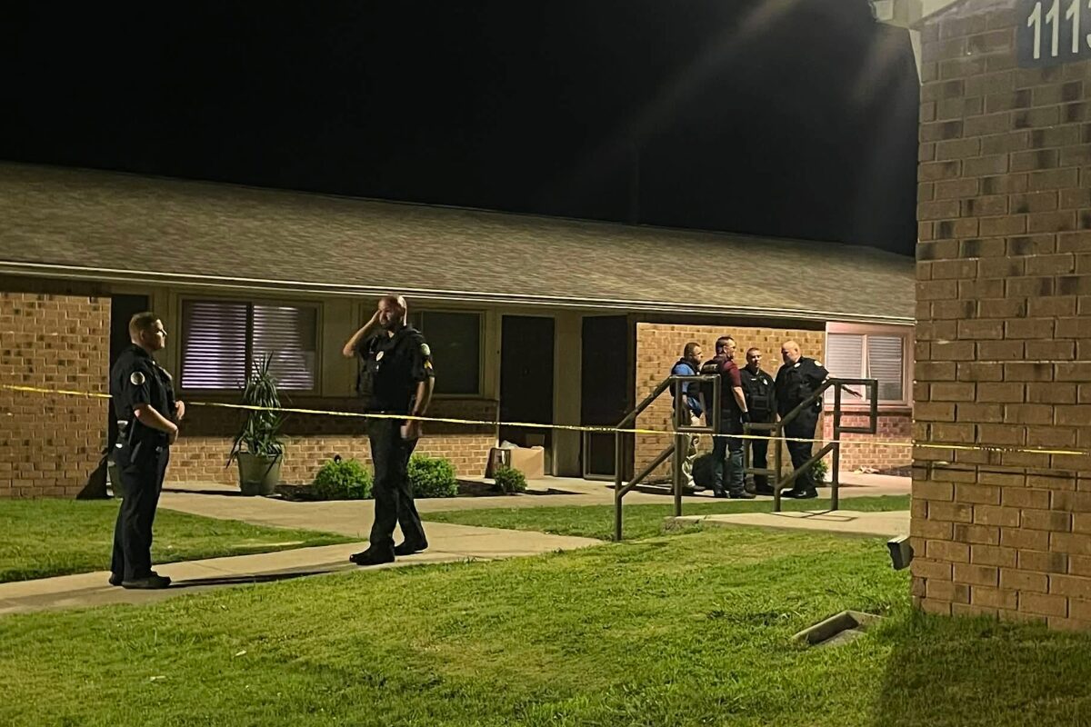 Police officers stand behind yellow caution tape outside a brick apartment building at night, investigating.