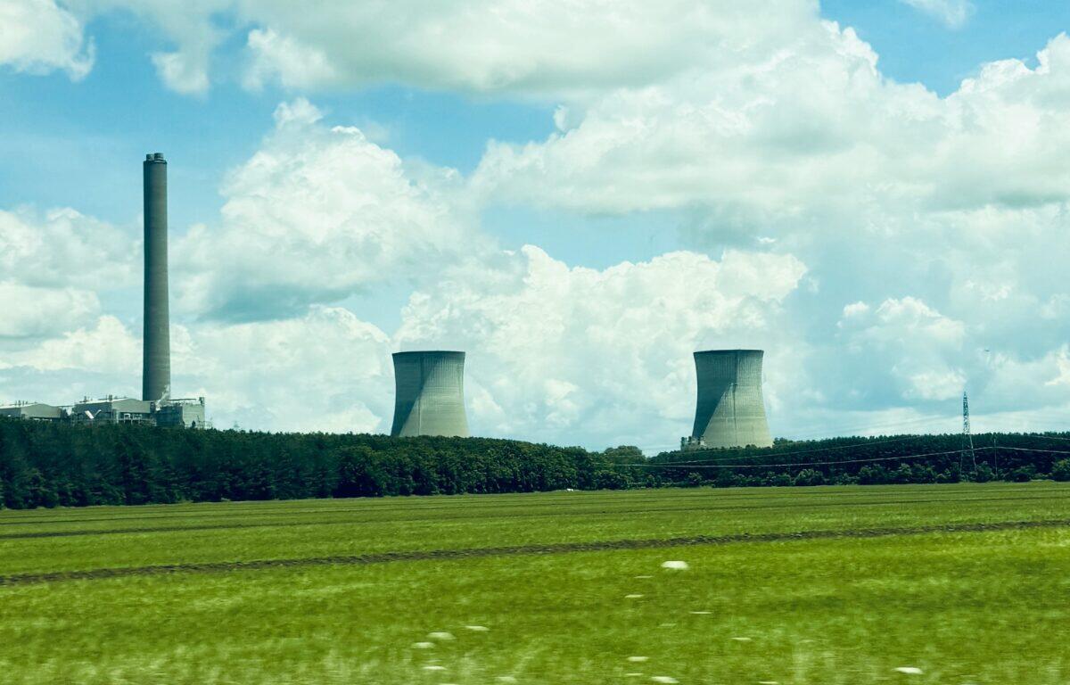 Industrial power plant with tall smokestack and two cooling towers over a green field under a blue sky with clouds.