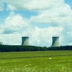 Industrial power plant with tall smokestack and two cooling towers over a green field under a blue sky with clouds.