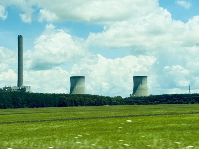 Industrial power plant with tall smokestack and two cooling towers over a green field under a blue sky with clouds.