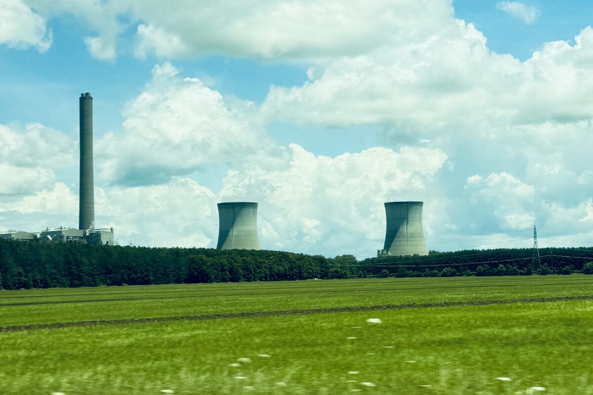 Industrial power plant with tall smokestack and two cooling towers over a green field under a blue sky with clouds.