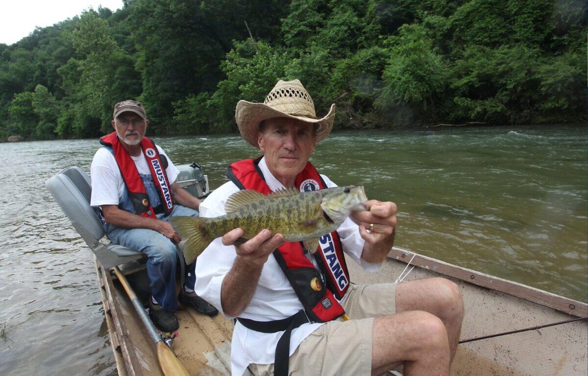 Two men in a small boat on a river, one wearing a straw hat and red life jacket holding a largemouth bass toward the camera while the other man sits behind him.
