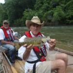 Two men in a small boat on a river, one wearing a straw hat and red life jacket holding a largemouth bass toward the camera while the other man sits behind him.