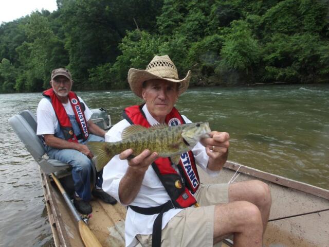 Two men in a small boat on a river, one wearing a straw hat and red life jacket holding a largemouth bass toward the camera while the other man sits behind him.