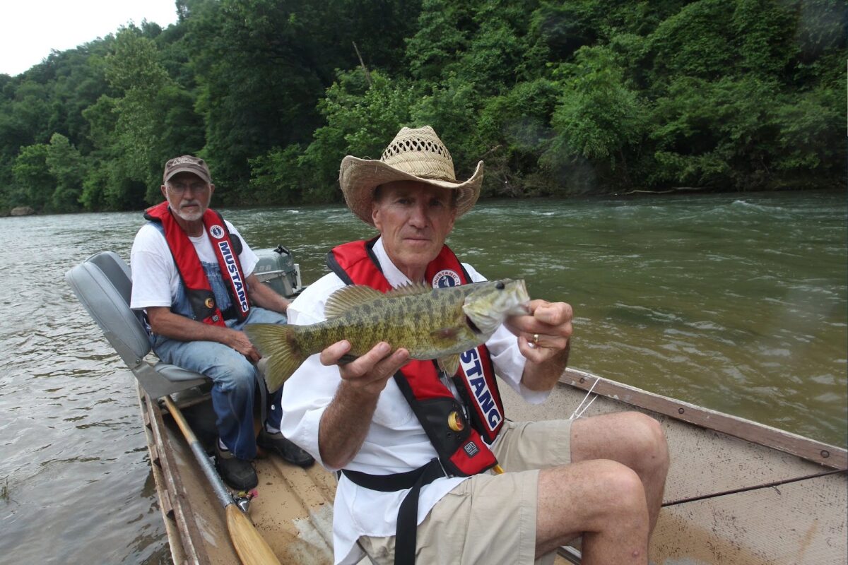 Two men in a small boat on a river, one wearing a straw hat and red life jacket holding a largemouth bass toward the camera while the other man sits behind him.