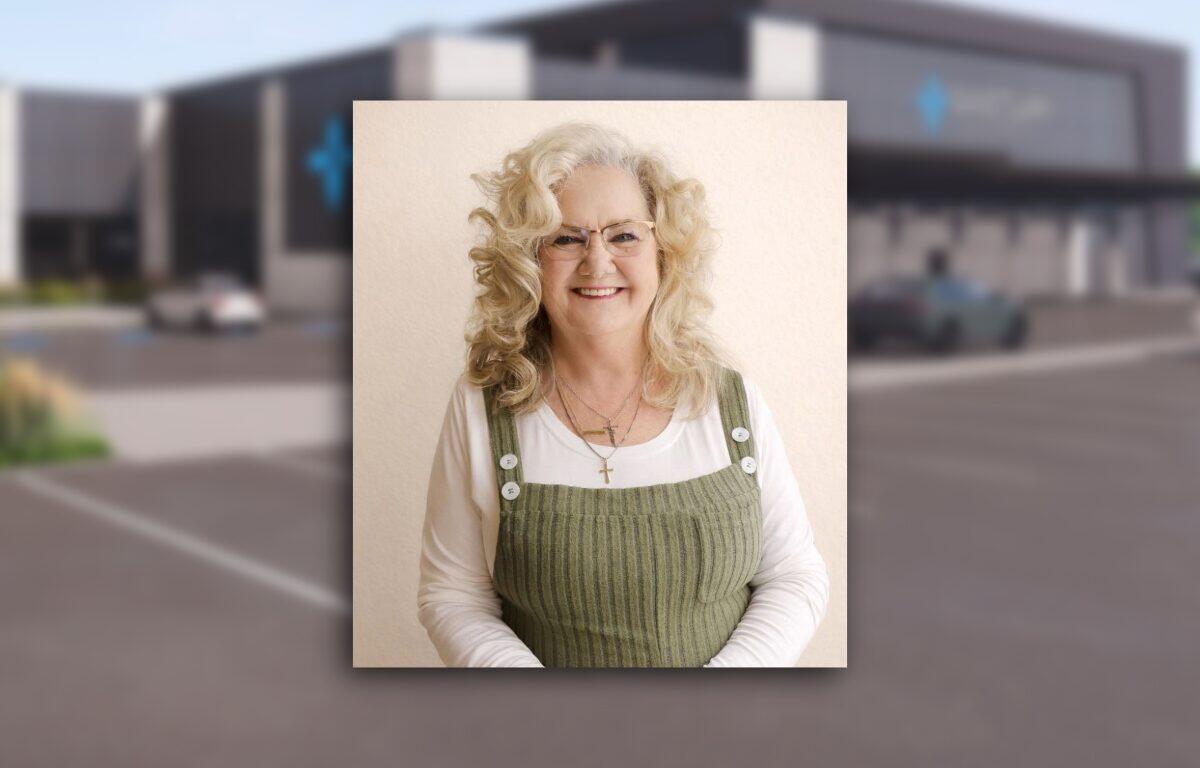 Portrait of a smiling older woman with curly blonde hair and glasses, wearing a white shirt under a green corduroy pinafore with white buttons.