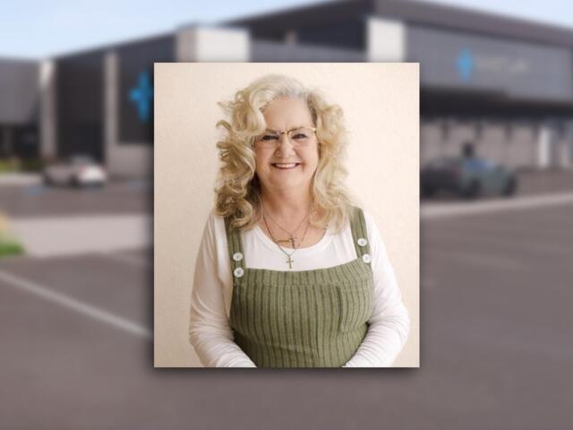 Portrait of a smiling older woman with curly blonde hair and glasses, wearing a white shirt under a green corduroy pinafore with white buttons.