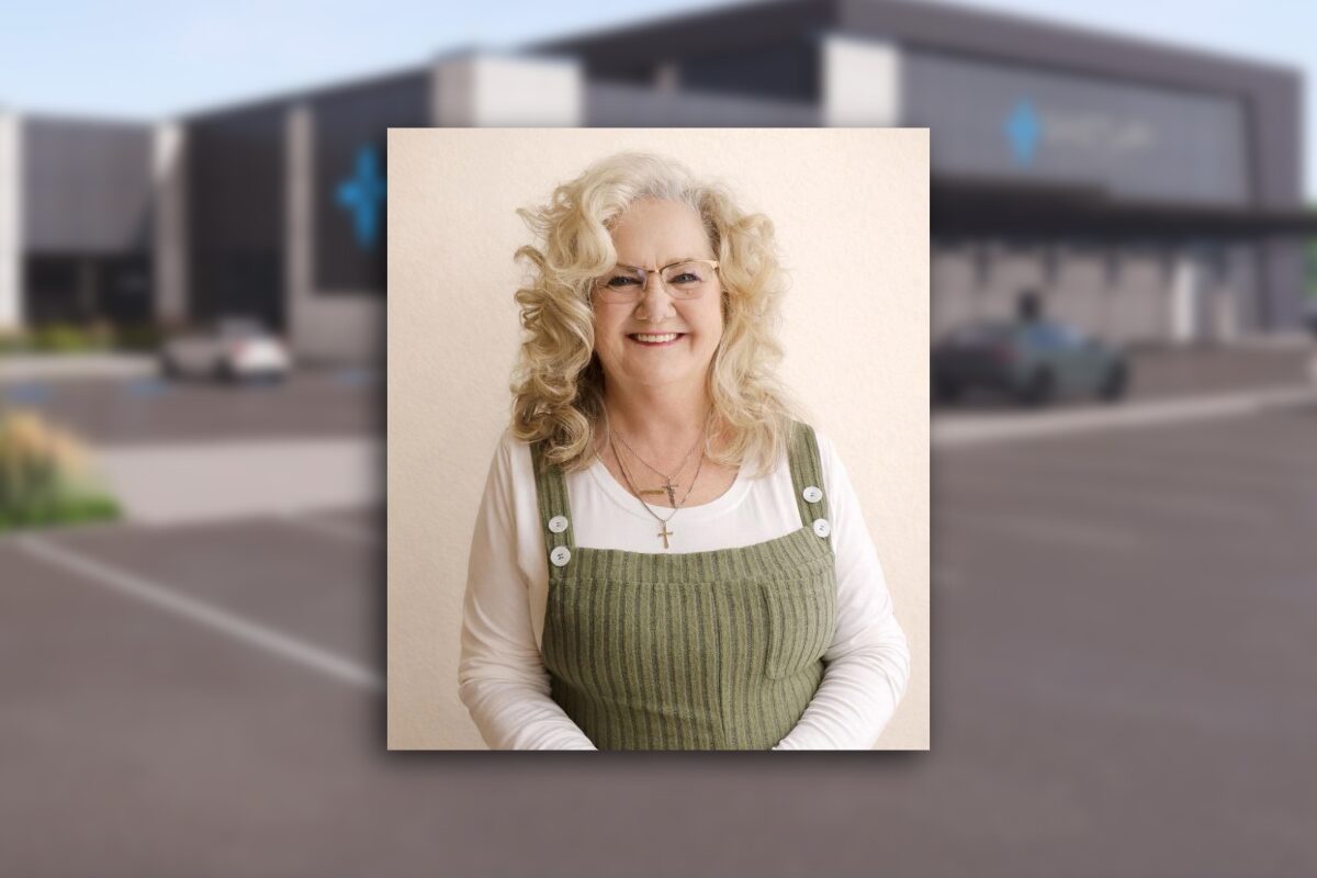 Portrait of a smiling older woman with curly blonde hair and glasses, wearing a white shirt under a green corduroy pinafore with white buttons.