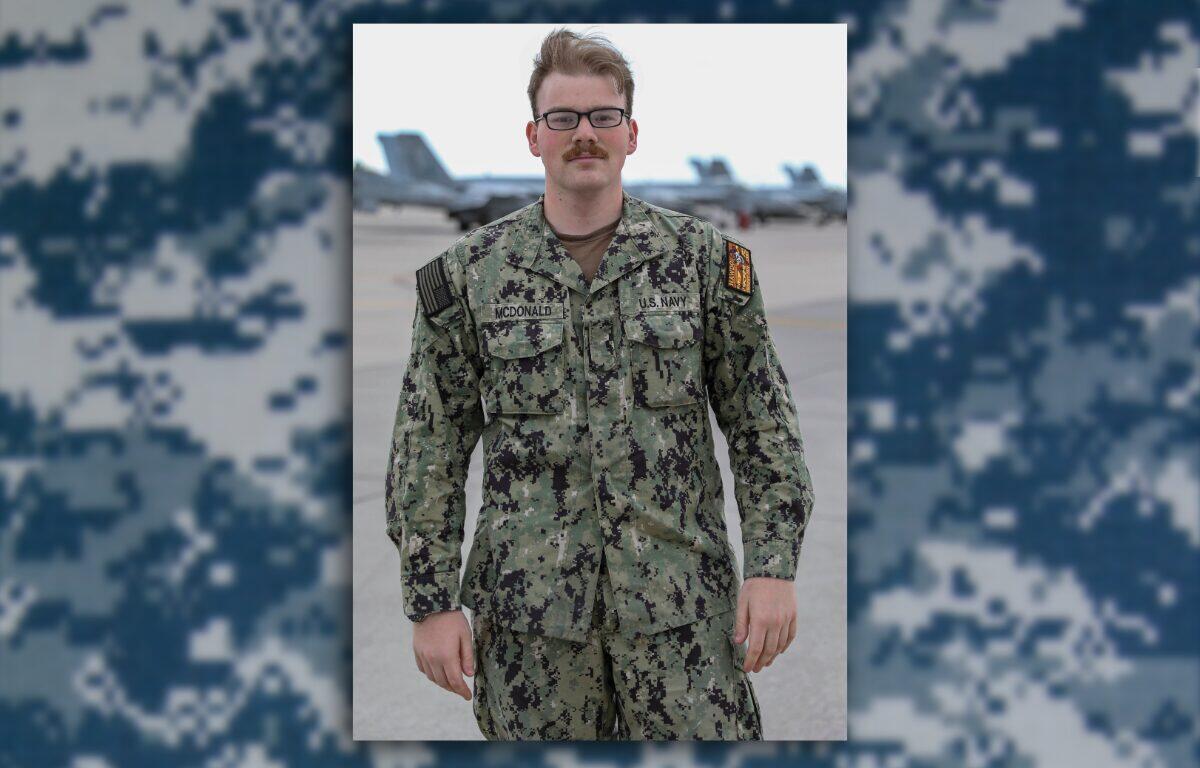 Male U.S. Navy service member in camouflage uniform standing on an airfield with jets in the background, name tag McDonald visible.