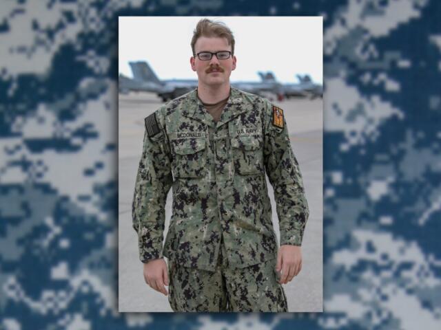 Male U.S. Navy service member in camouflage uniform standing on an airfield with jets in the background, name tag McDonald visible.