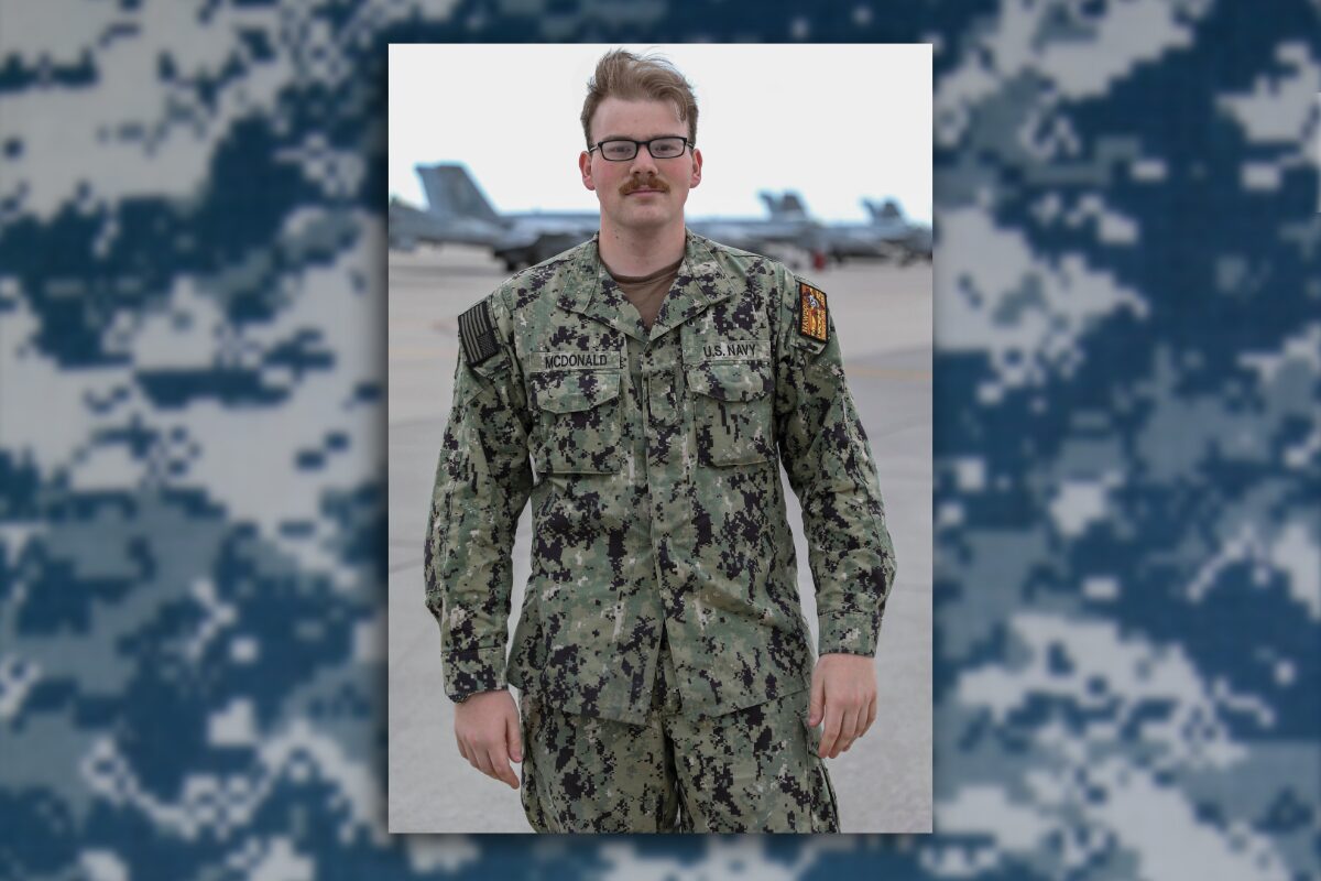 Male U.S. Navy service member in camouflage uniform standing on an airfield with jets in the background, name tag McDonald visible.
