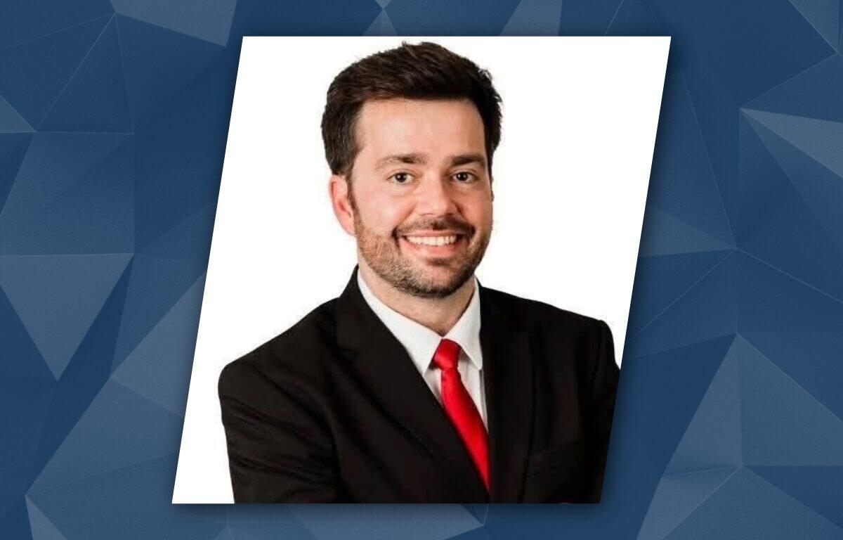Smiling man in a black suit and red tie, head-and-shoulders portrait for a professional profile against a white background (slightly tilted).