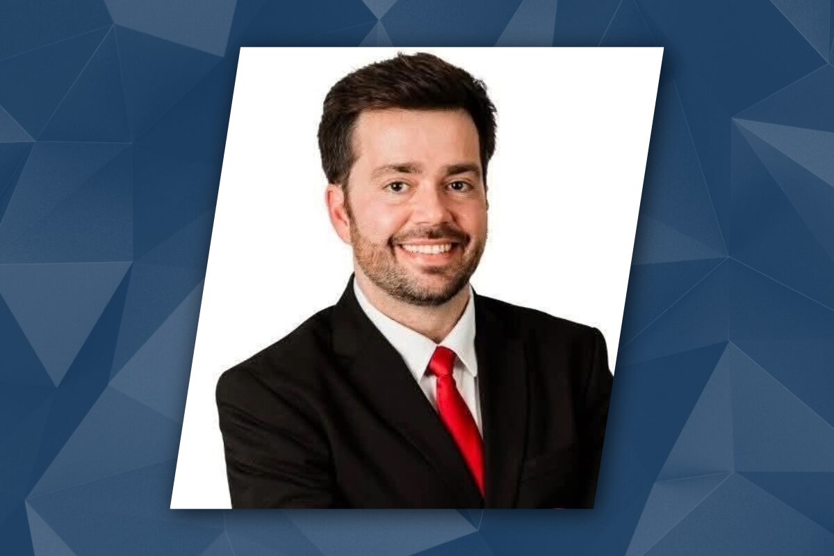 Smiling man in a black suit and red tie, head-and-shoulders portrait for a professional profile against a white background (slightly tilted).