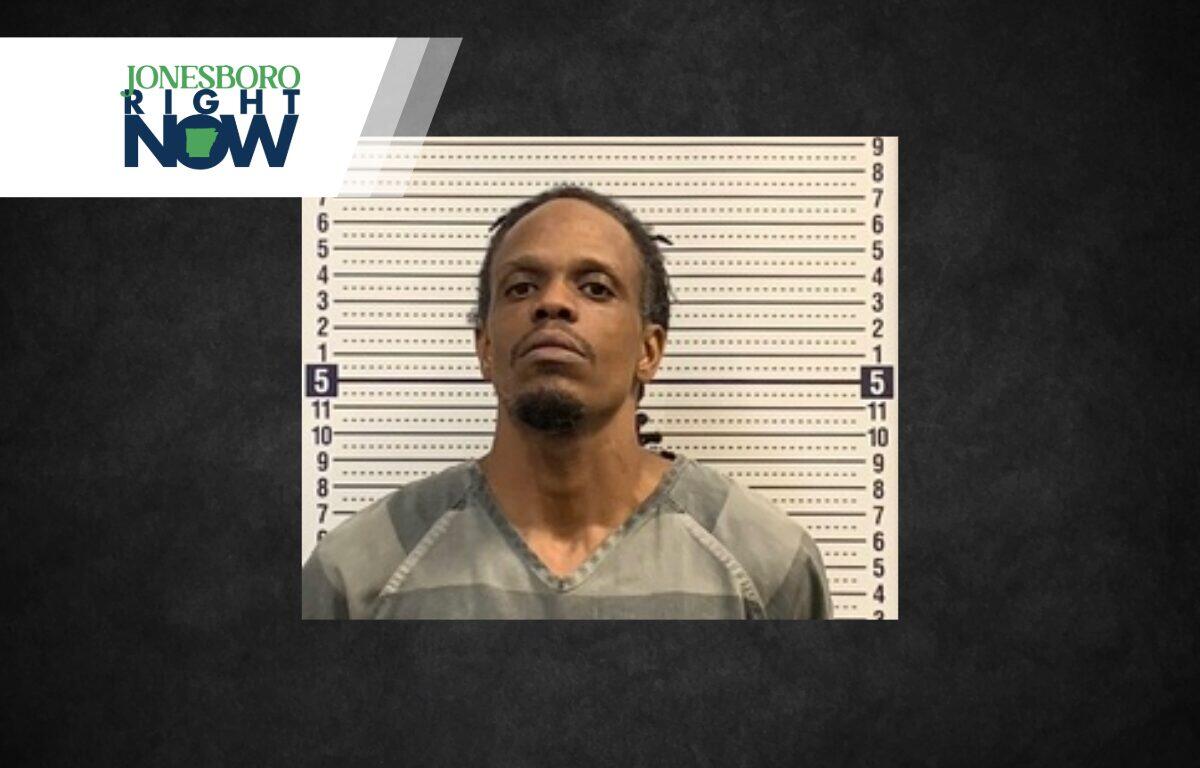 Mugshot of a man in a gray inmate jumpsuit standing before a height chart, with Jonesboro Now logo in the top-left corner.