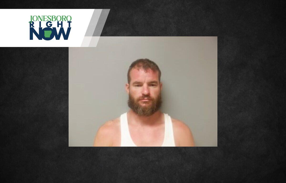Mugshot-style photo of a bearded man in a white tank top, standing against a plain grey wall, with a Jonesboro Right Now logo in the top-left corner.