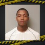 Mugshot of a young man with short hair wearing a white shirt, neutral expression, gray wall background, police tape border framing the image.