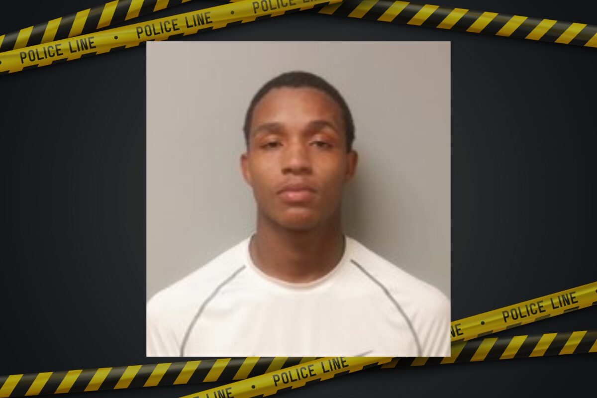 Mugshot of a young man with short hair wearing a white shirt, neutral expression, gray wall background, police tape border framing the image.