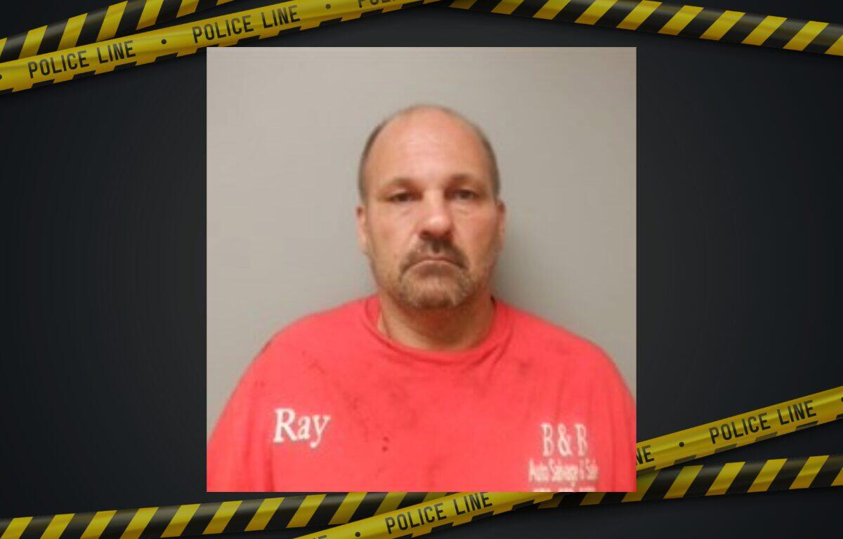 Mugshot of a middle-aged man in a red shirt with 'Ray' on the chest, gray background, framed by police tape