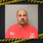 Mugshot of a middle-aged man in a red shirt with 'Ray' on the chest, gray background, framed by police tape