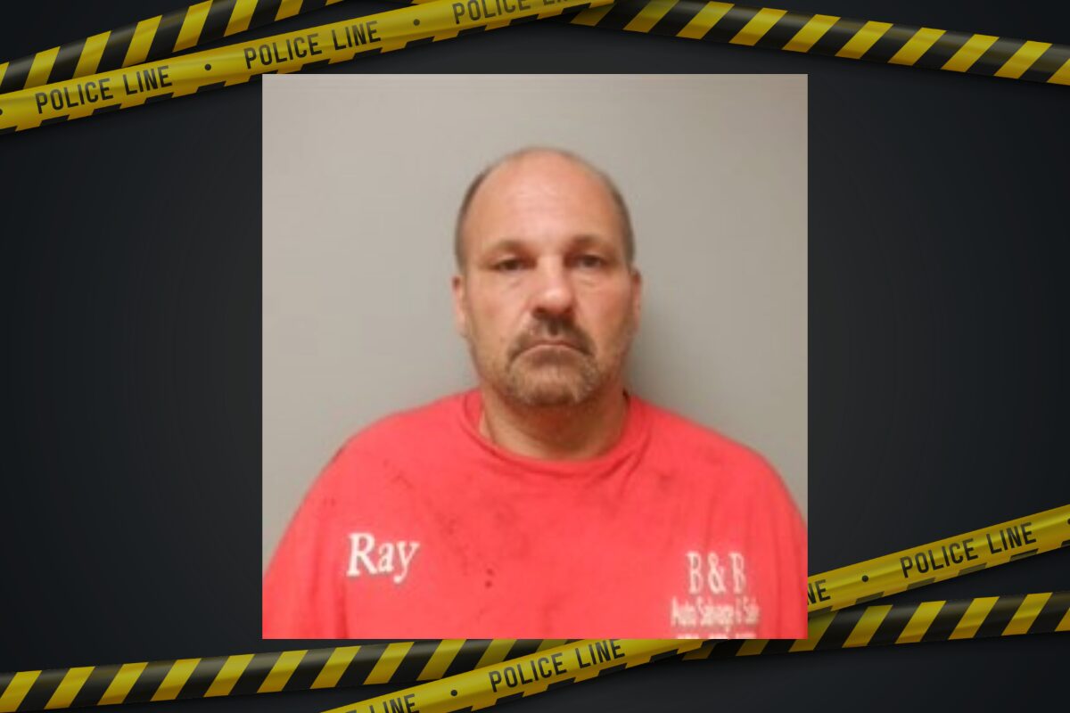 Mugshot of a middle-aged man in a red shirt with 'Ray' on the chest, gray background, framed by police tape