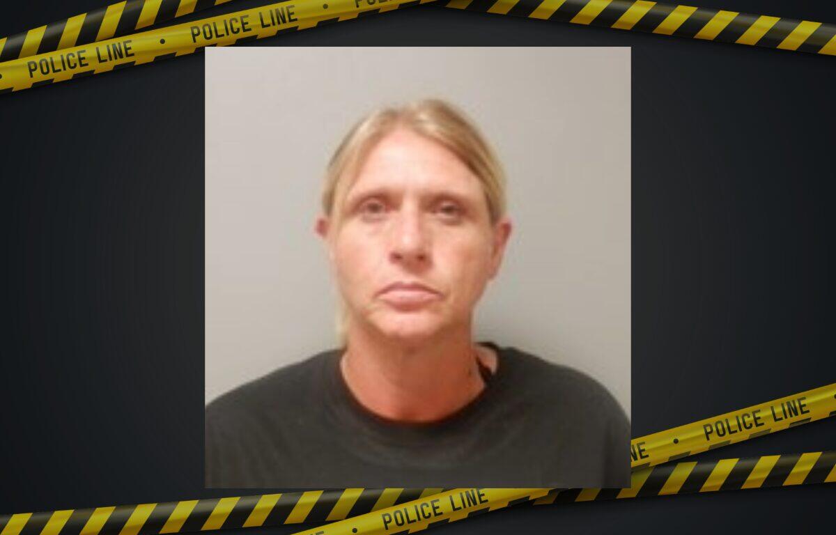 Mugshot of a blonde woman in a dark shirt, neutral expression, against a plain background with police tape framing the image.
