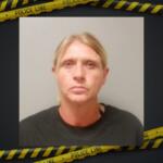 Mugshot of a blonde woman in a dark shirt, neutral expression, against a plain background with police tape framing the image.