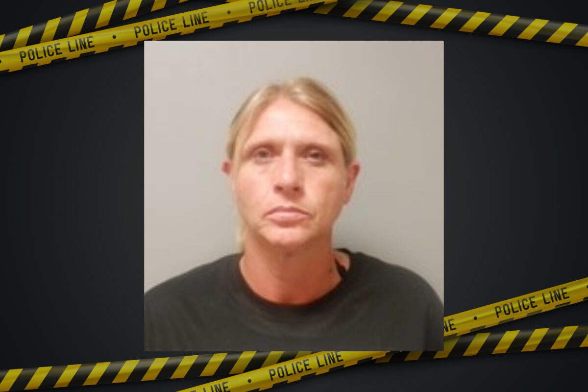 Mugshot of a blonde woman in a dark shirt, neutral expression, against a plain background with police tape framing the image.