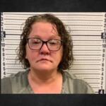 Mugshot of a middle-aged woman with glasses in a gray top, standing before a height-measure backdrop.